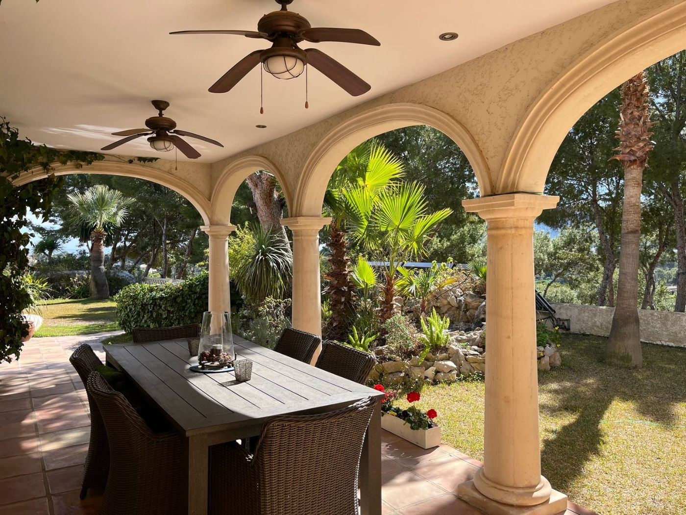 Terraza cubierta con arcos de piedra, suelos de terracota y comedor exterior frente a un jardín mediterráneo con palmeras y vegetación frondosa.