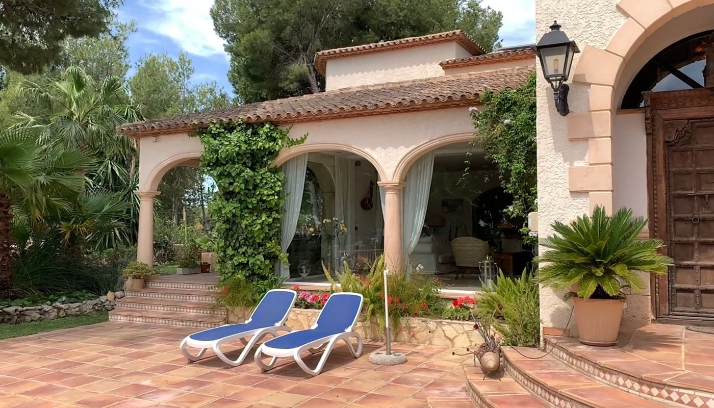 Terraza exterior con baldosas de terracota, tumbonas azules y porche de arcos clásicos junto a una puerta de madera tallada y jardín mediterráneo.