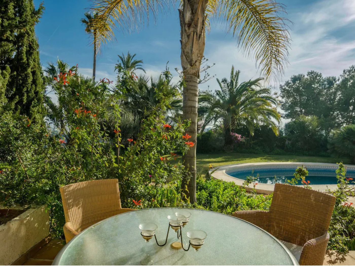 Terraza exterior con mesa de cristal y asientos de mimbre, con vistas a una piscina circular y jardines mediterráneos con palmeras y vegetación densa.