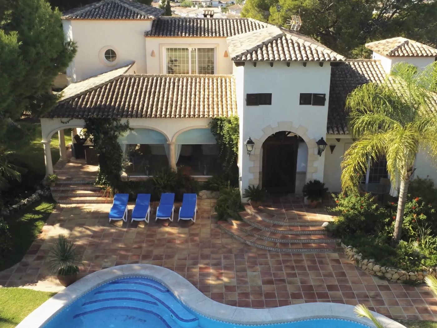 Vista elevada de una villa mediterránea con piscina curva, terraza de terracota con cuatro hamacas y fachada blanca con entradas de piedra en arco.