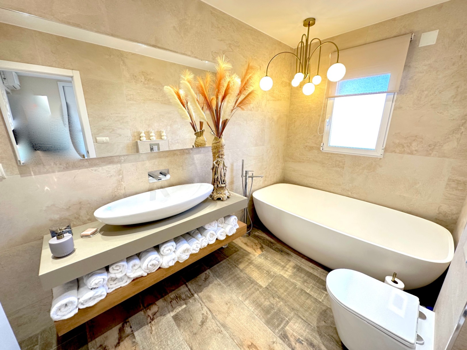 Baño contemporáneo con bañera exenta, lavabo sobre encimera y sanitario suspendido. Destaca la iluminación de diseño y el revestimiento cerámico en tonos arena.