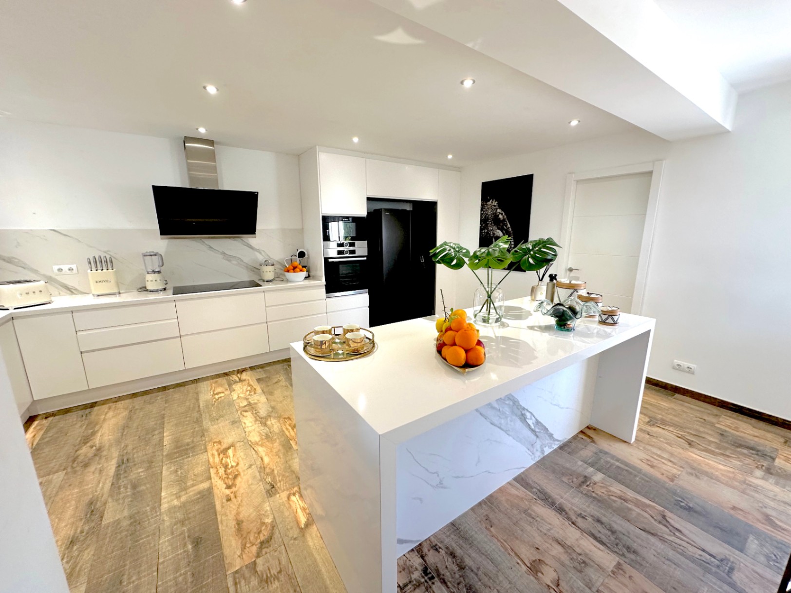 Cocina de diseño contemporáneo con isla central de efecto mármol, mobiliario blanco minimalista, electrodomésticos integrados en negro y suelos de gres porcelánico imitación madera.