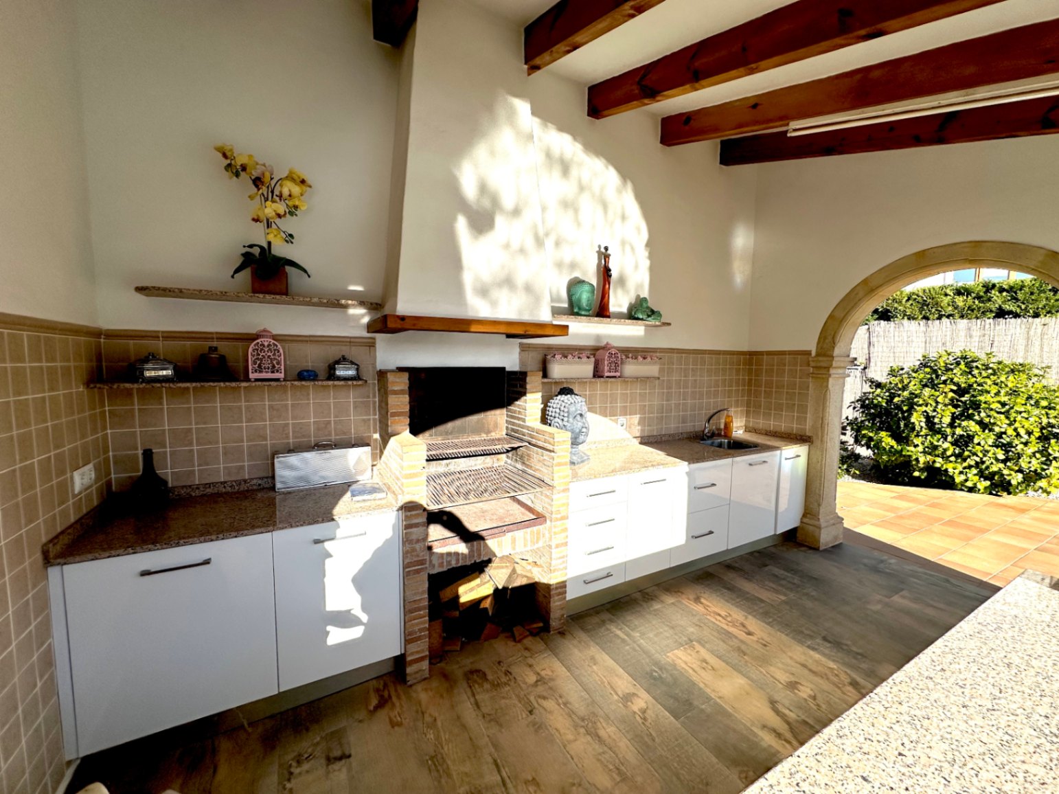 Cocina de verano con barbacoa de obra, encimeras de granito y vigas de madera. Incluye zona de fregadero y acceso exterior mediante un arco de piedra tradicional.