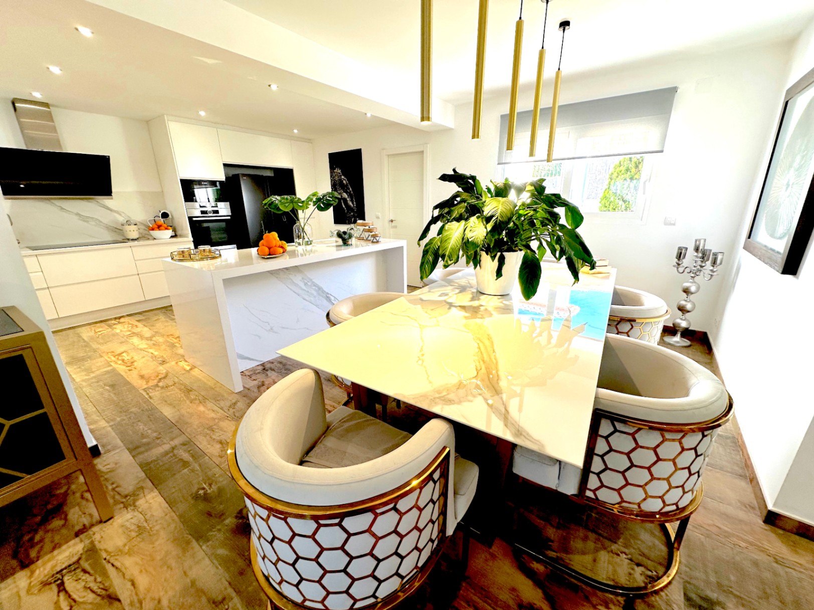 Comedor de concepto abierto con mesa de mármol y sillas de diseño dorado, integrado con una cocina moderna de isla central, acabados marmoleados y suelos de gres cerámico.