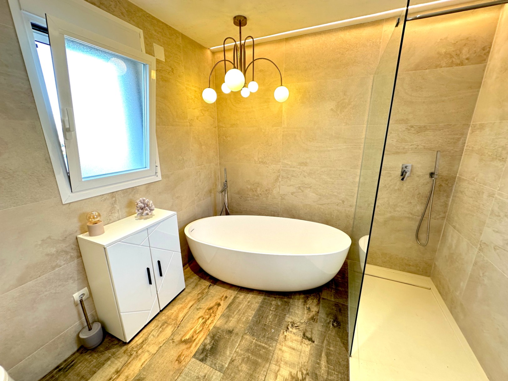 Cuarto de baño con bañera exenta ovalada, ducha con mampara de cristal, suelos de gres efecto madera y paredes revestidas en piedra natural.