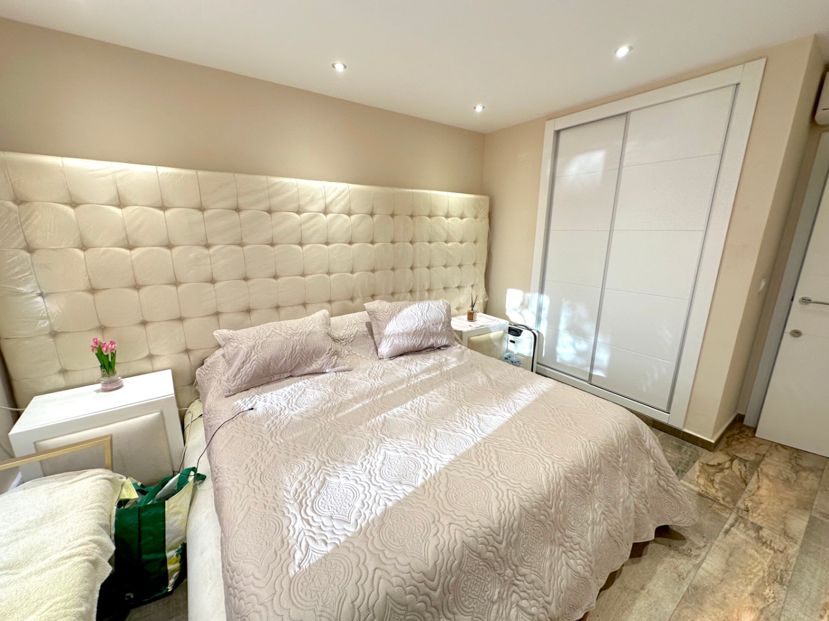 Dormitorio con un gran cabecero capitoné, iluminación empotrada en el techo y un armario empotrado con puertas correderas blancas sobre suelos de piedra natural.