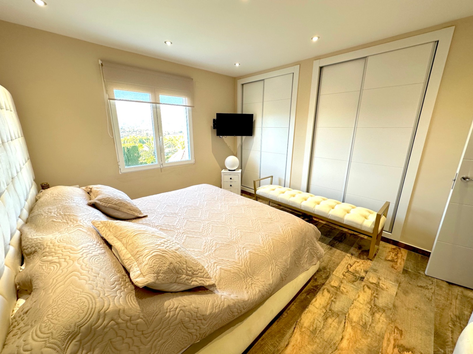 Dormitorio luminoso con armarios empotrados de puertas correderas, suelo cerámico de imitación madera, iluminación empotrada y ventana con vistas despejadas al entorno natural.