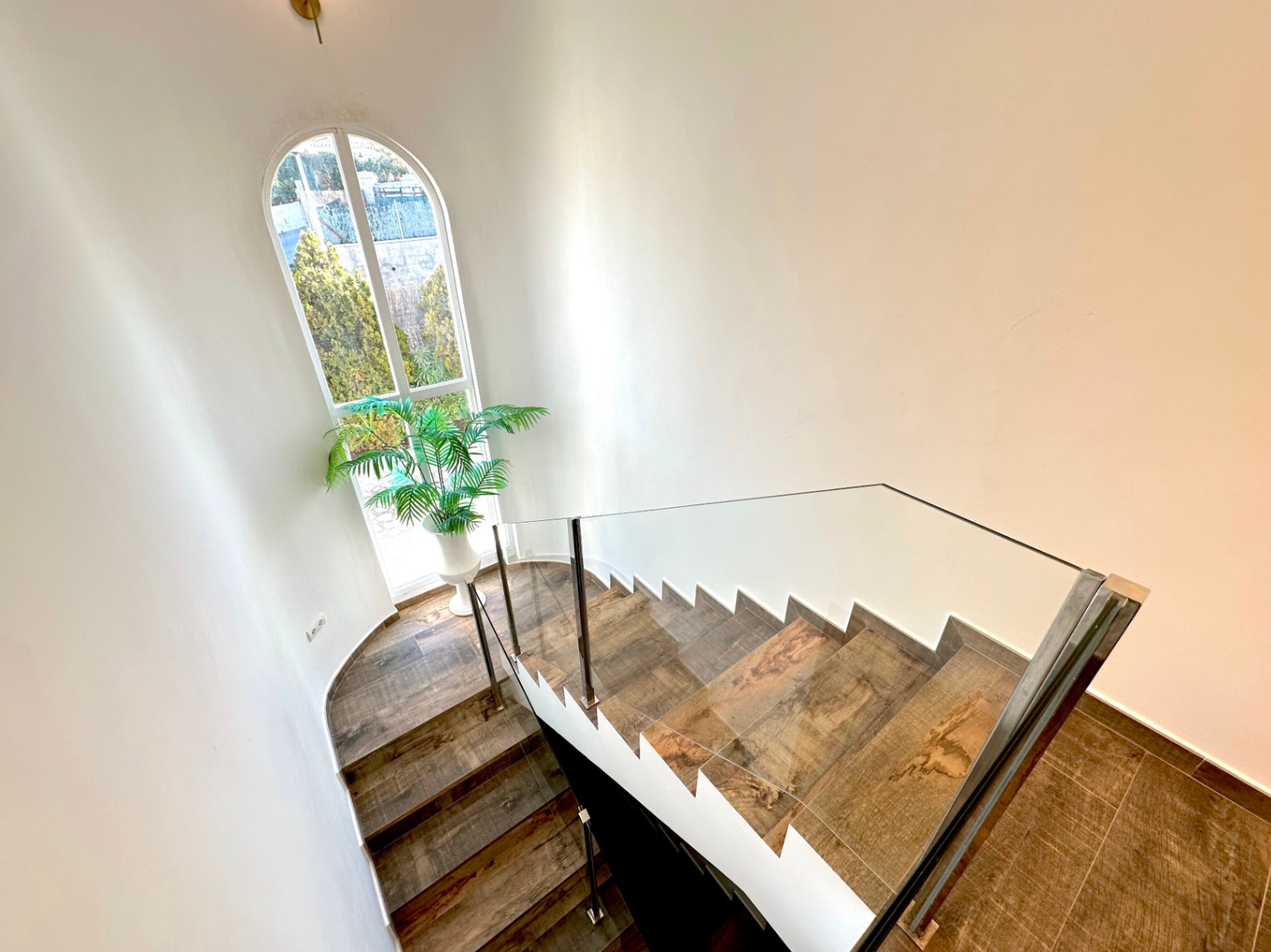 Escalera con peldaños de gres efecto madera, barandilla de cristal y un ventanal de medio punto que ilumina el espacio con vistas al jardín.