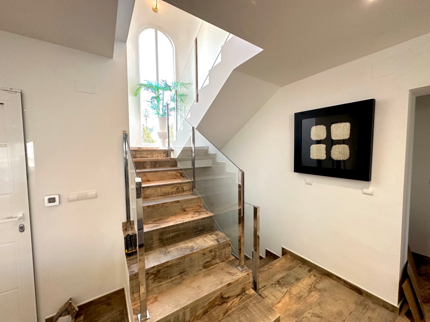 Escalera interior con peldaños de efecto madera, barandilla de cristal y acero inoxidable, y un gran ventanal de arco que aporta luz natural.