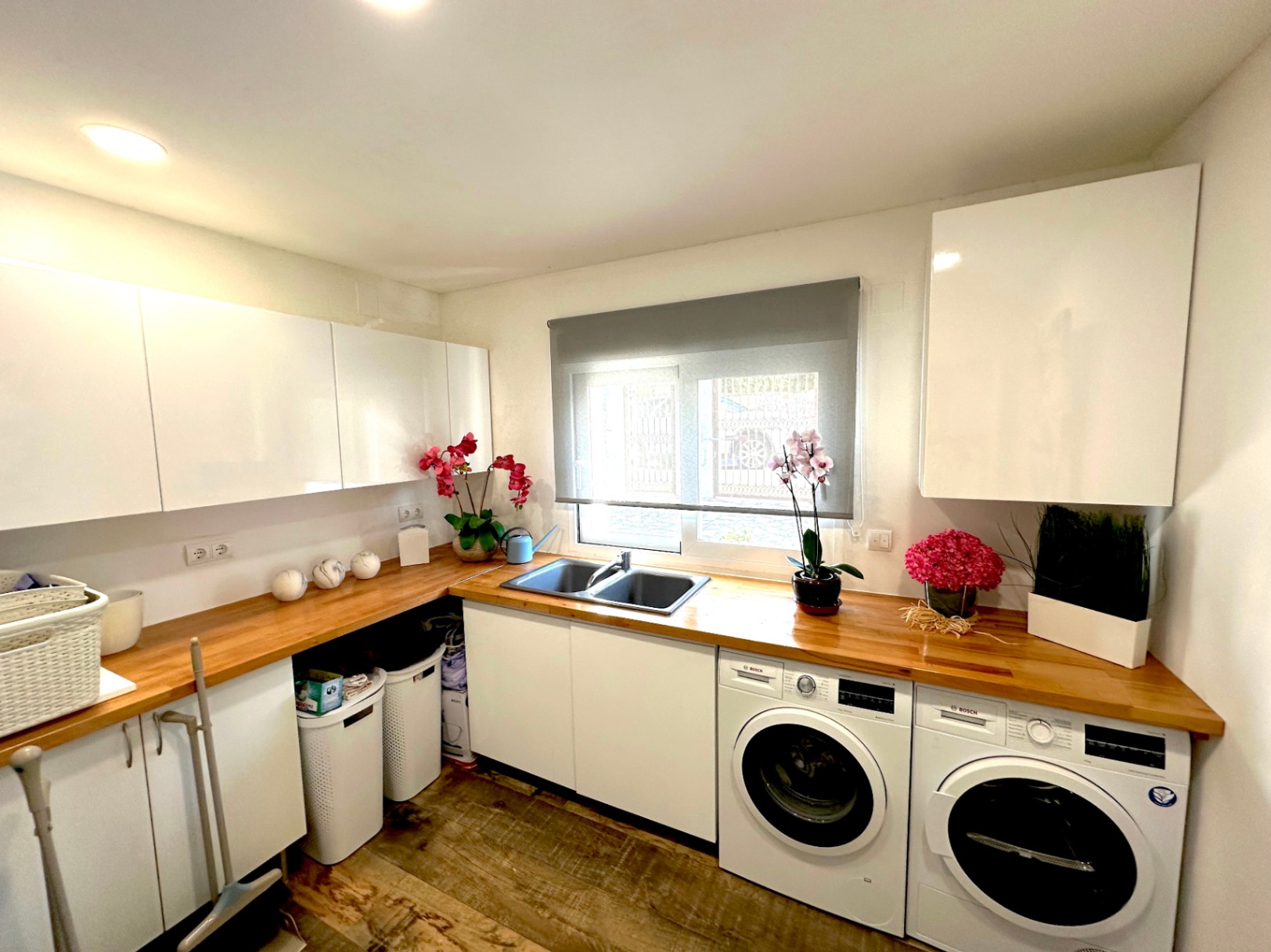Lavadero funcional con encimera de madera natural, armarios blancos de alto brillo y electrodomésticos Bosch integrados bajo una ventana con estor.