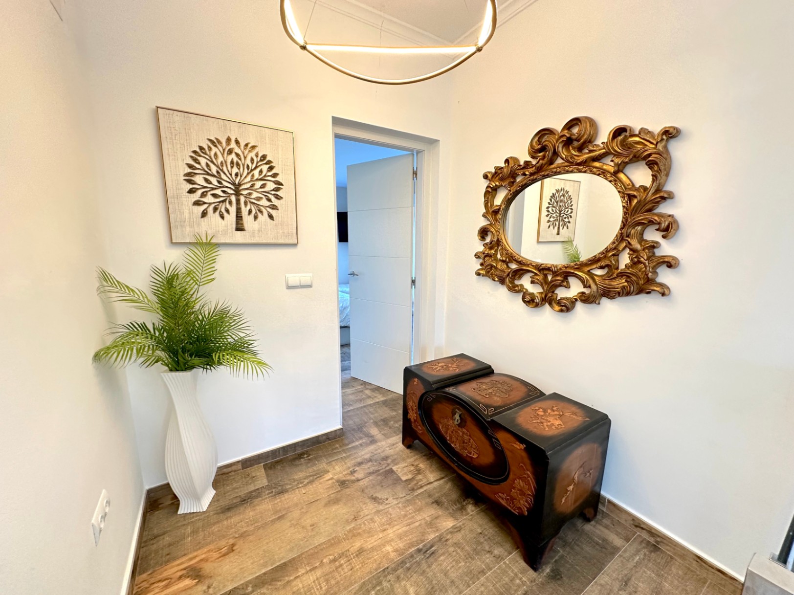 Pasillo con suelos de gres efecto madera, un baúl tallado de estilo clásico y un espejo con marco dorado ornamental bajo una iluminación LED circular moderna.