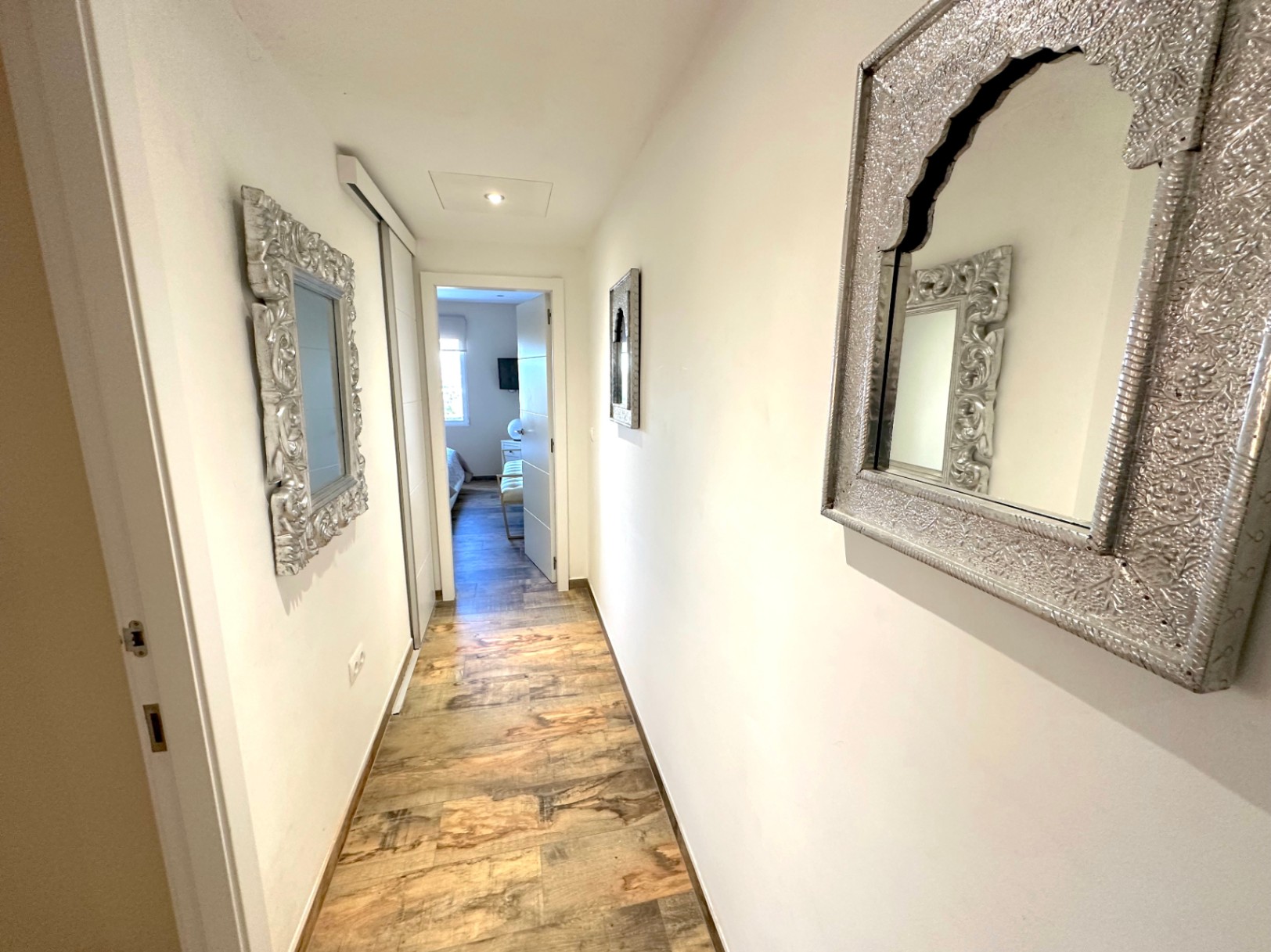 Pasillo luminoso con suelos de gres efecto madera, espejos de marco plateado ornamentado y acceso a dormitorios con armarios empotrados integrados.