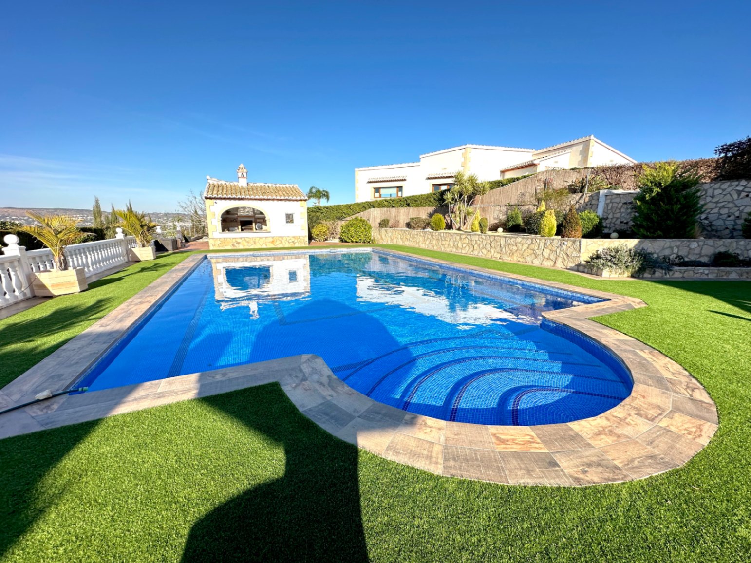 Piscina exterior con escalera romana, rodeada de césped artificial y muros de piedra natural. Incluye una cocina de verano independiente y vistas despejadas hacia el valle.