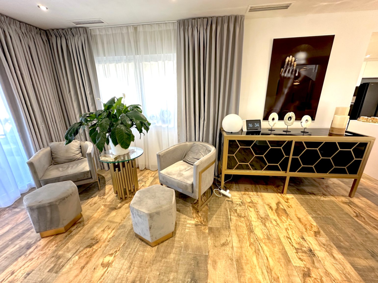 Rincón de descanso con sillones de terciopelo gris, pufs hexagonales y aparador de diseño con detalles geométricos dorados sobre suelo de madera.