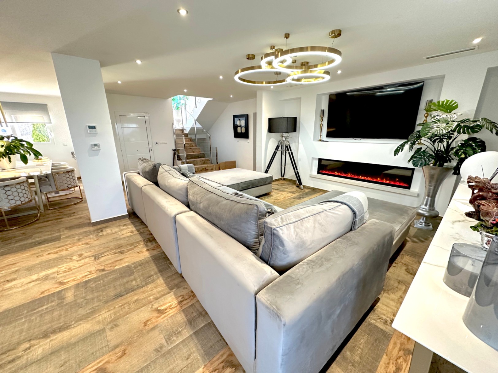 Salón de concepto abierto con sofá modular gris, chimenea lineal integrada bajo televisor y lámparas circulares de diseño. Suelos de gres imitación madera y escalera con barandilla de cristal.