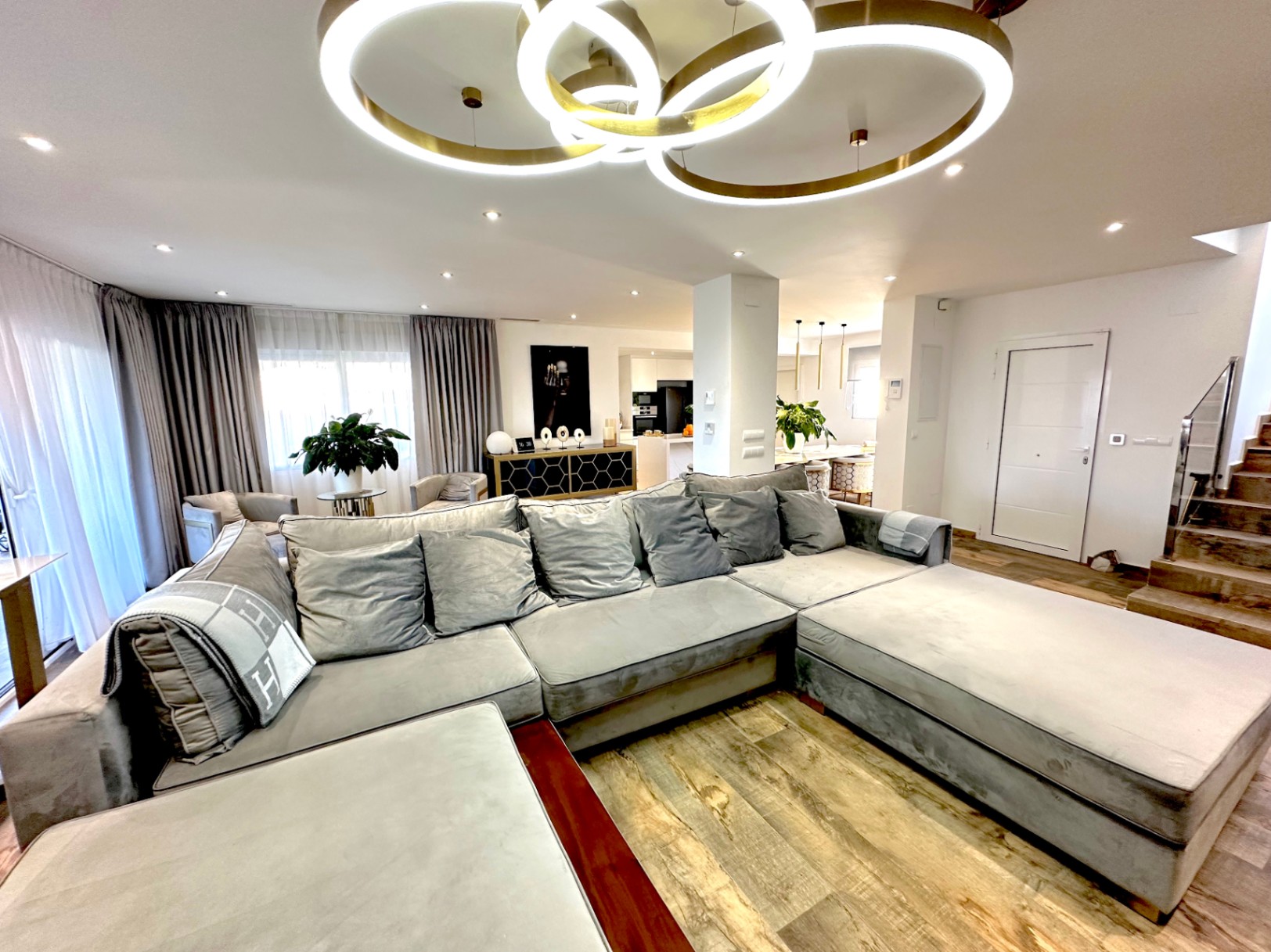 Salón de concepto abierto con un amplio sofá modular gris, suelos de gres efecto madera y una moderna lámpara de techo de anillos LED dorados.