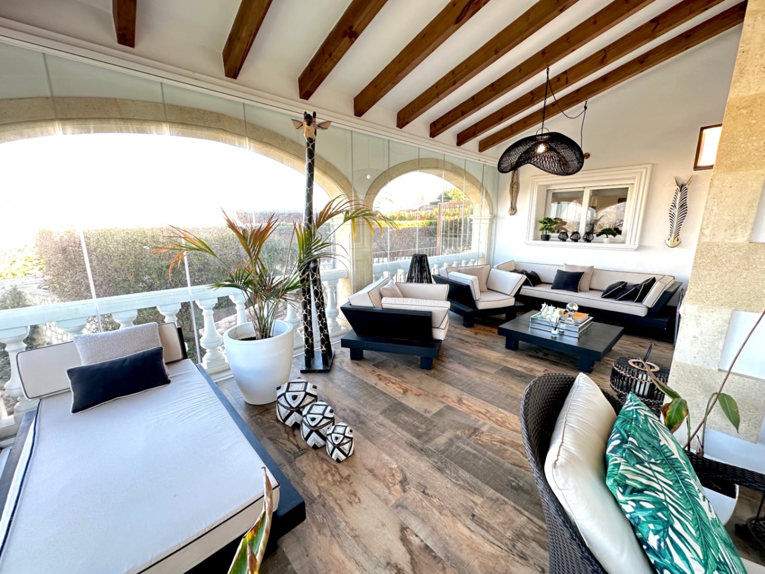 Terraza acristalada con vigas de madera vista, mobiliario contemporáneo en blanco y negro y arcos tradicionales con vistas al jardín.