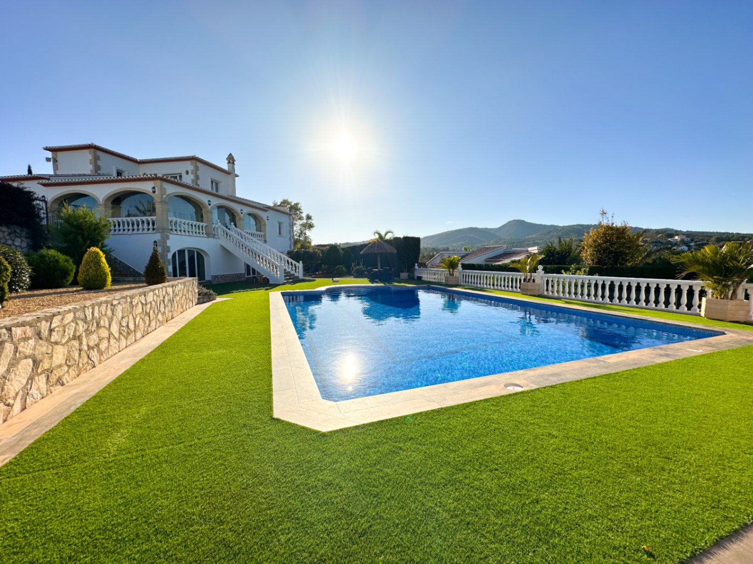 Vista exterior de la piscina rectangular rodeada de césped artificial, con la villa blanca de arcos tradicionales y muros de piedra natural al fondo.