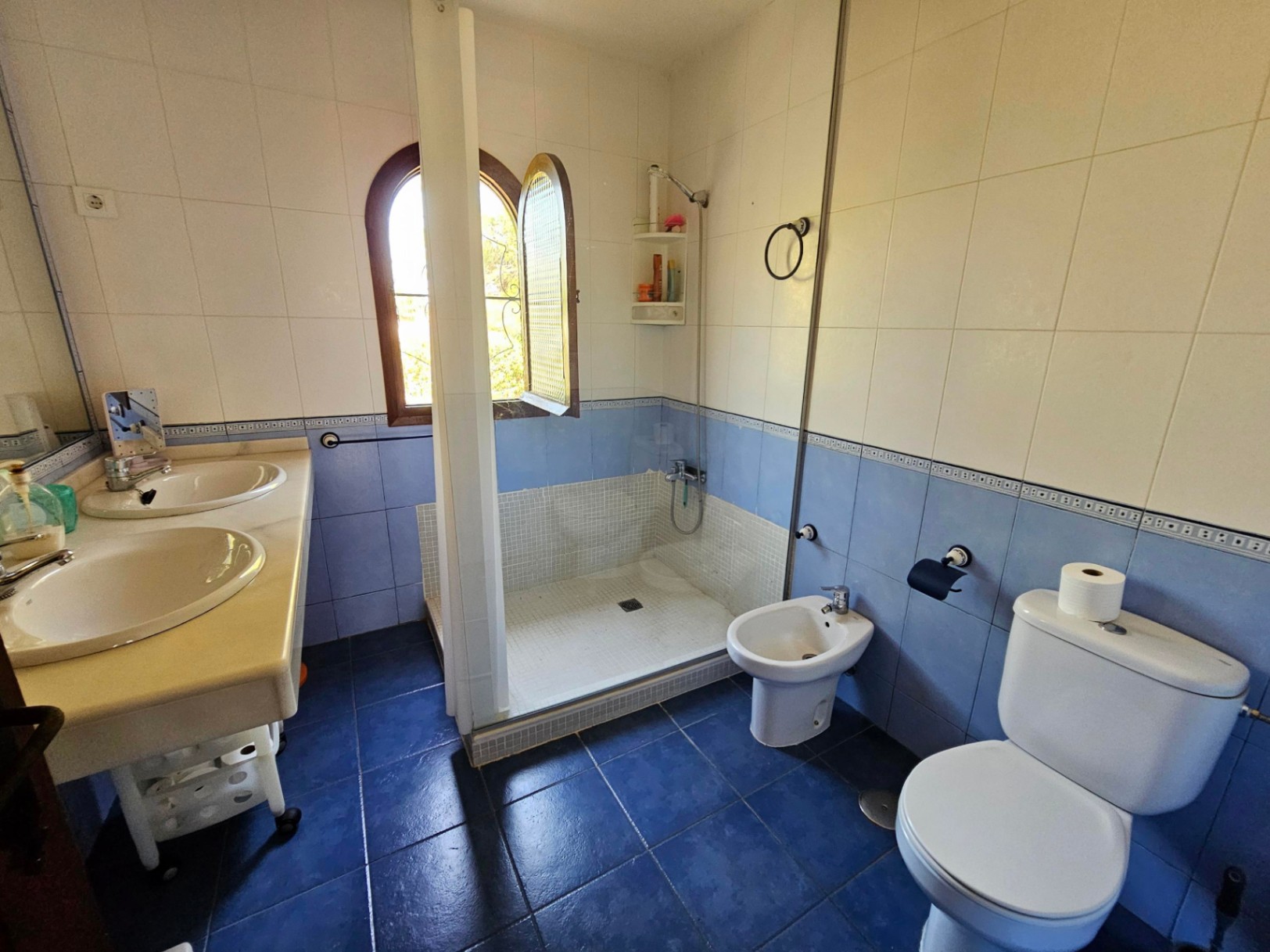 Baño con lavabo doble, ducha de mosaico y ventana de arco. Paredes y suelos revestidos con azulejos en tonos azules y blancos.