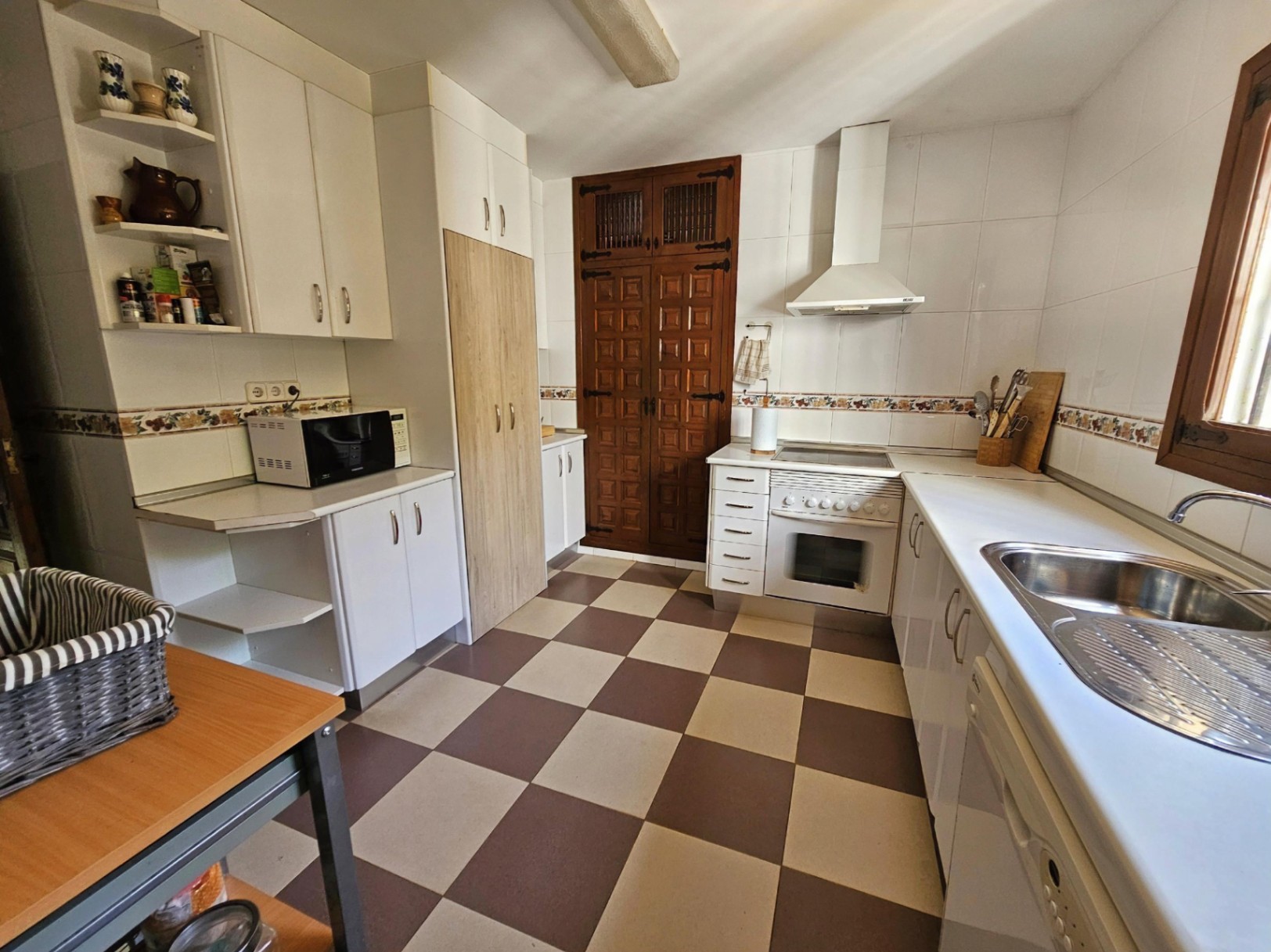 Cocina con suelo de baldosas damero, armarios blancos y una puerta de despensa de madera rústica tallada. Incluye encimera blanca y fregadero de acero inoxidable.