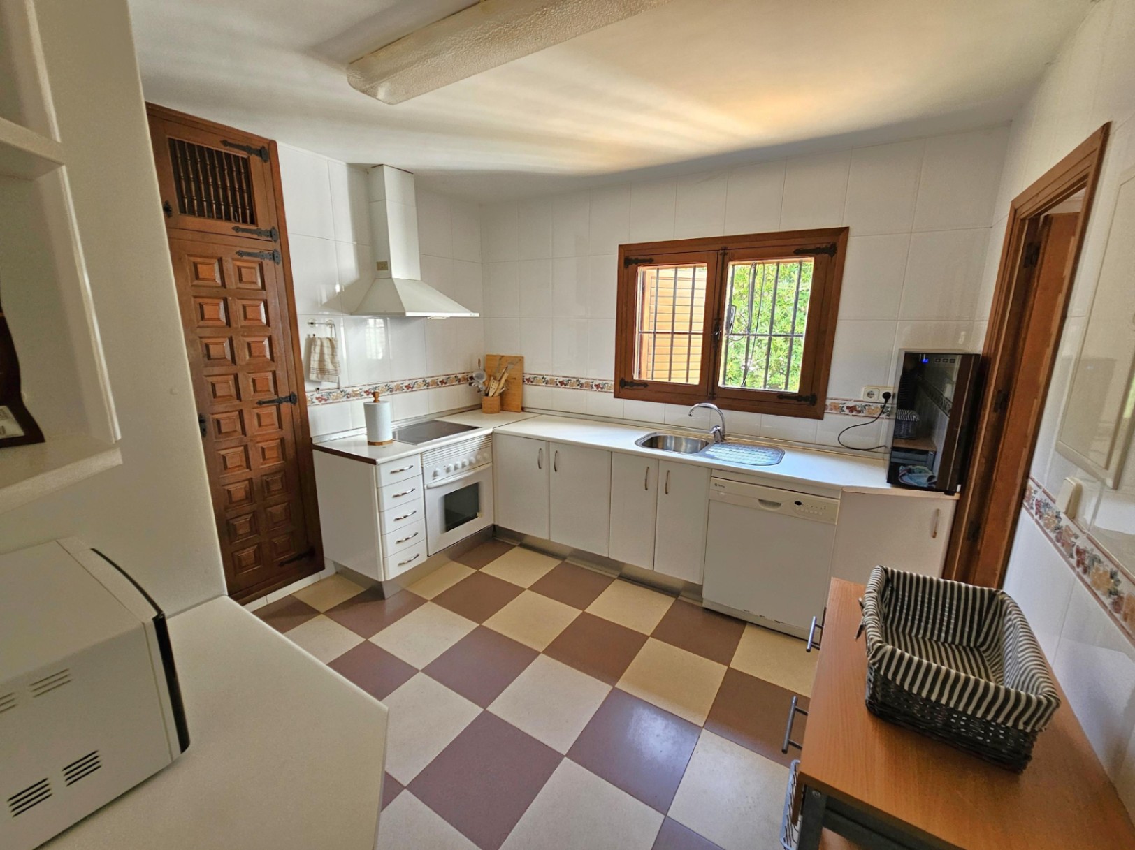 Cocina con suelo de baldosas en damero, armarios blancos y una puerta de despensa de madera tallada con herrajes de hierro tradicionales.