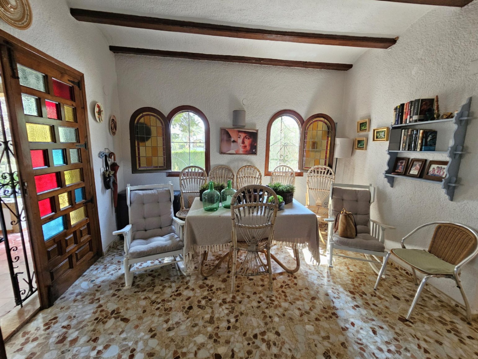 Comedor con suelo de terrazo, vigas de madera a la vista y ventanas de arco. Destaca la puerta de madera con cristales de colores y el mobiliario de mimbre.