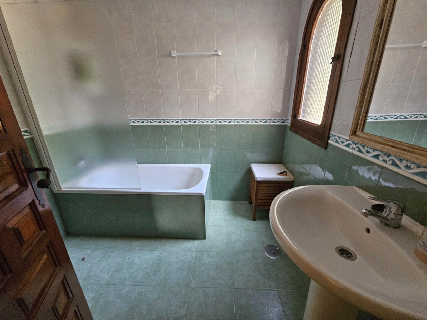 Cuarto de baño con bañera integrada, mampara de cristal esmerilado, azulejos verdes con cenefa decorativa y ventana de madera en arco.