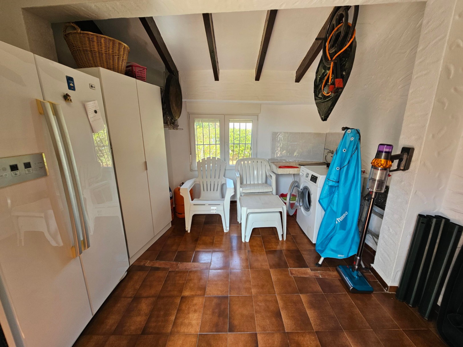 Cuarto de servicio con suelo de baldosas cerámicas, vigas de madera vista, lavadora y armarios blancos bajo un techo de estilo rústico.