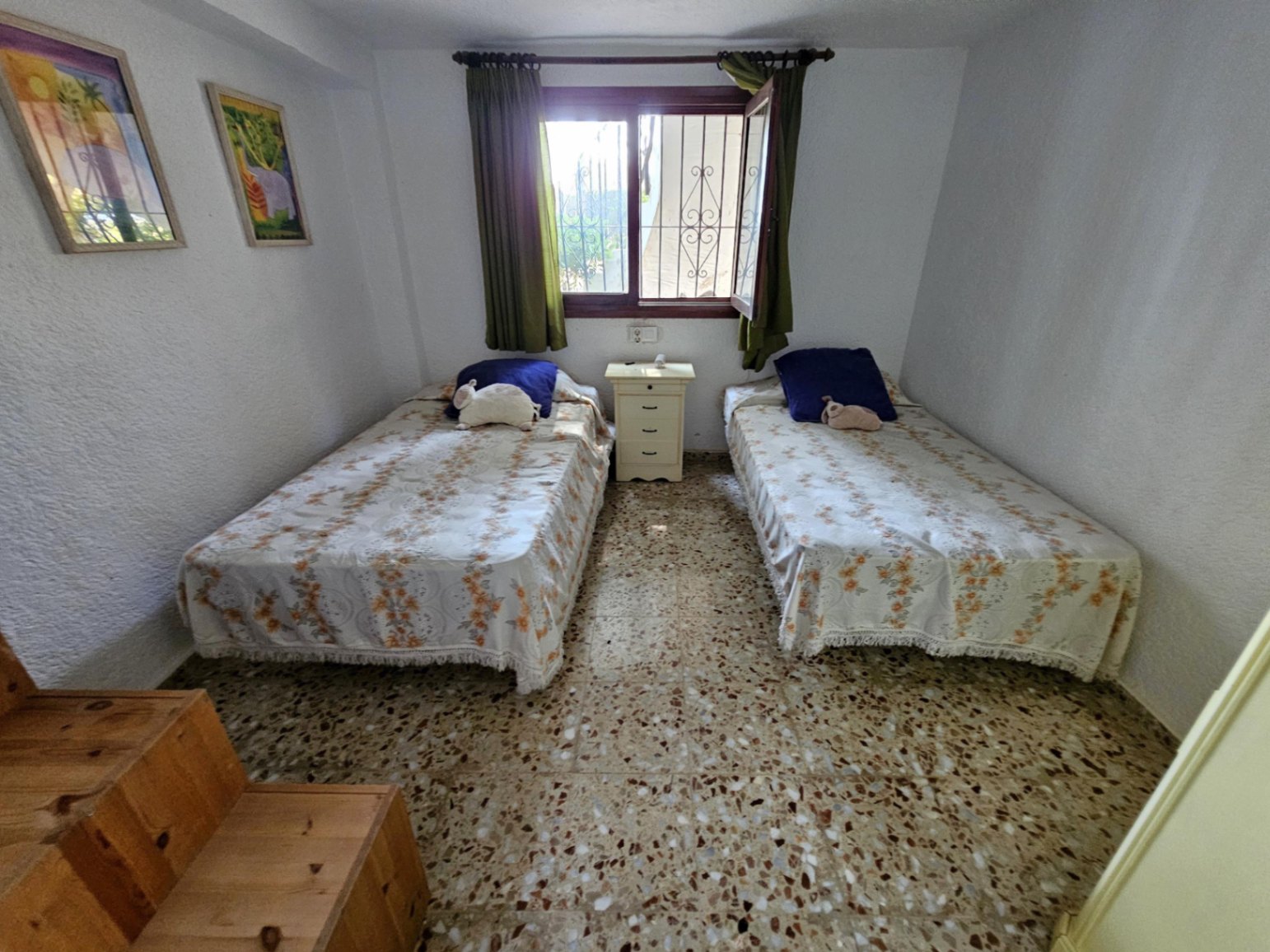 Dormitorio con dos camas individuales, suelo de terrazo tradicional y ventana central con rejas y cortinas verdes. Incluye una mesita de noche blanca entre las camas.