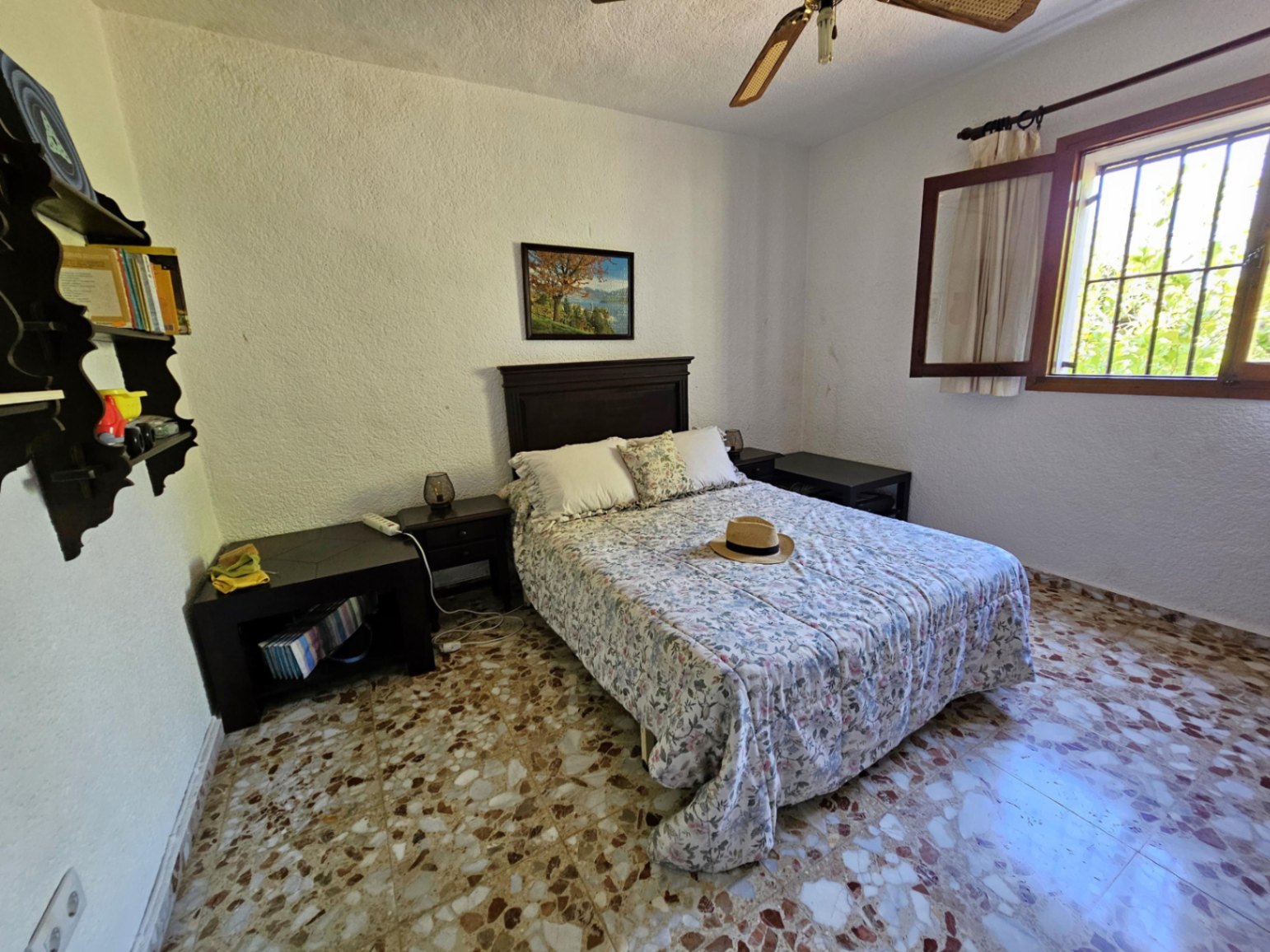 Dormitorio con suelos de terrazo original, cama doble con cabecero de madera oscura y ventana con marco de madera que permite la entrada de luz natural.