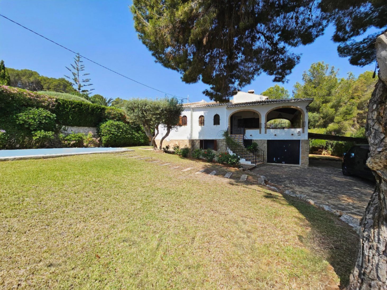 Exterior de una villa mediterránea blanca con porche de arcos, base de piedra natural, amplio jardín de césped y zona de piscina rodeada de pinos maduros.
