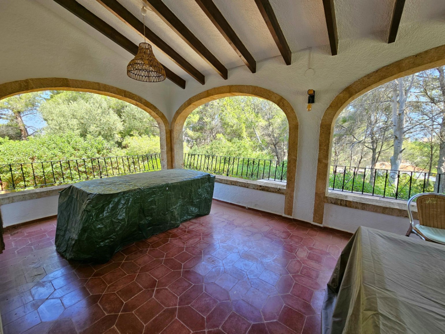 Terraza cubierta con vigas de madera vistas, suelo de baldosas hexagonales de terracota y arcos de piedra con vistas al bosque.