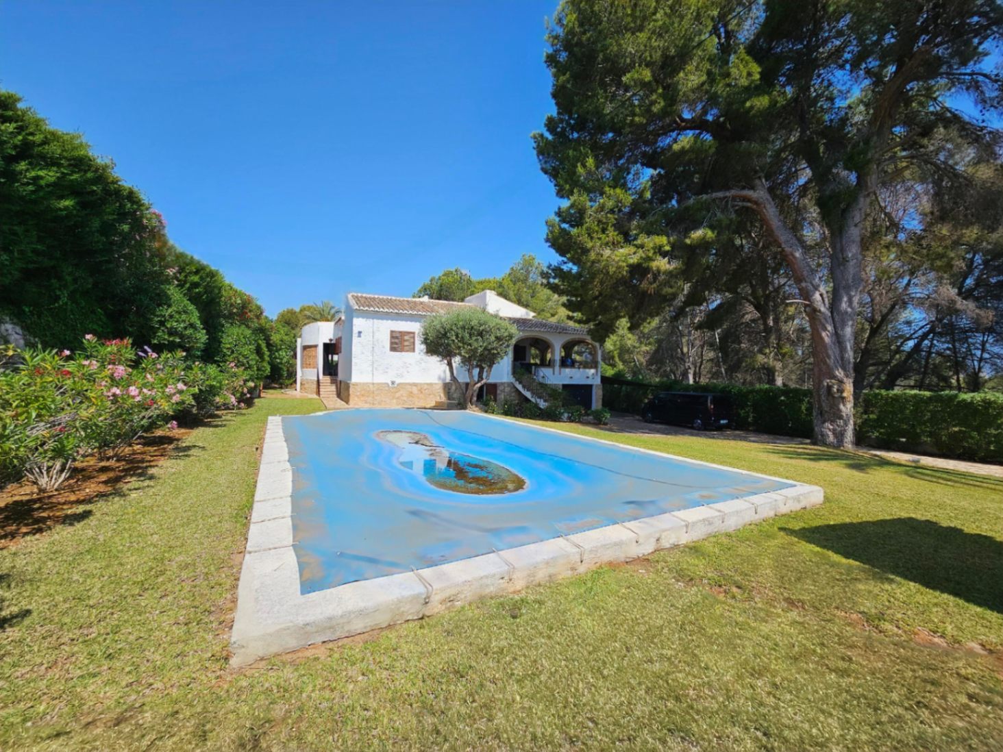 Villa mediterránea de fachada blanca y porche con arcos, con piscina rectangular protegida por una cubierta azul, rodeada de jardín con césped y pinos maduros.