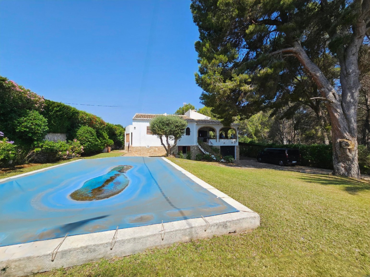 Vista exterior de una villa blanca con terraza de arcos y amplio jardín de césped con piscina cubierta, rodeada de pinos maduros y vegetación mediterránea.