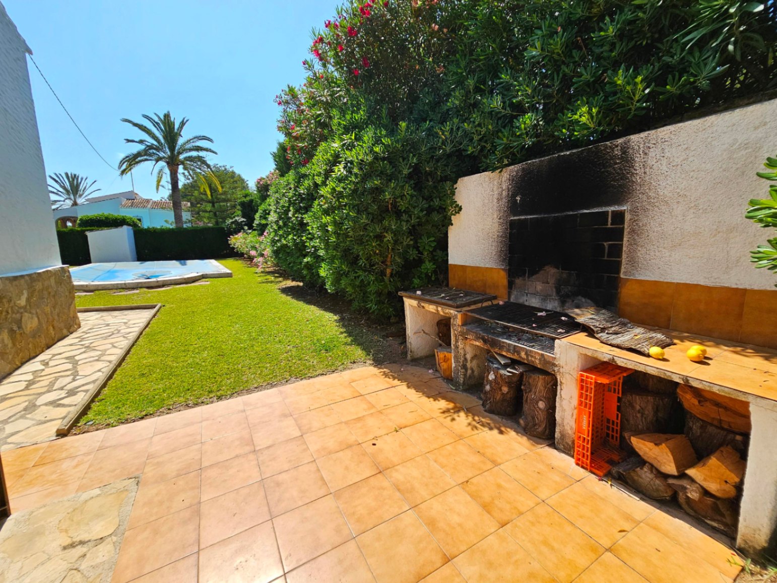Zona de barbacoa de obra con encimeras cerámicas y almacenamiento de leña, situada junto a un jardín de césped y una piscina rodeada de vegetación mediterránea.