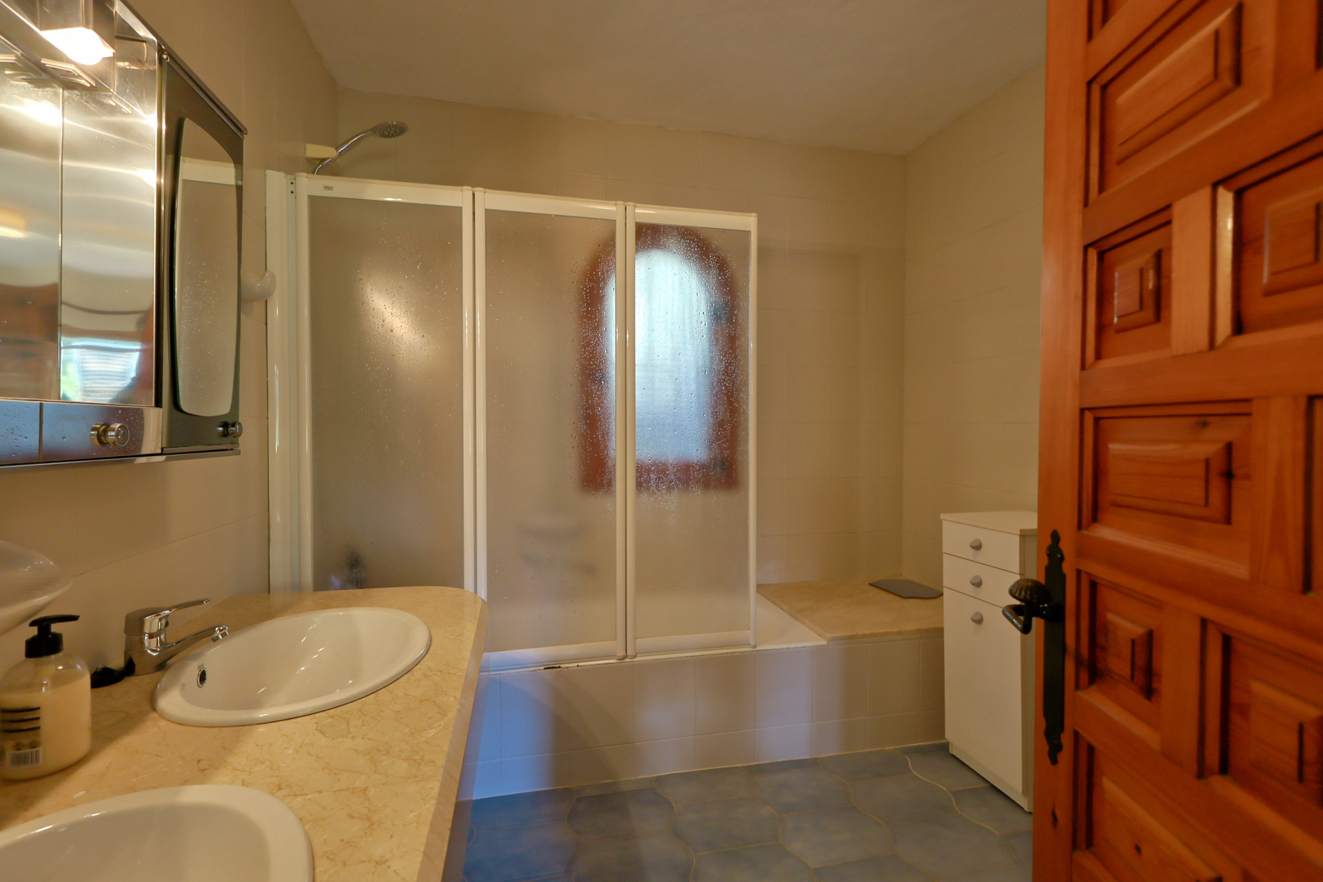 Baño con encimera de mármol de doble seno, armario de espejo y bañera con mampara plegable translúcida. Suelo de baldosas azules y puerta de madera tallada.