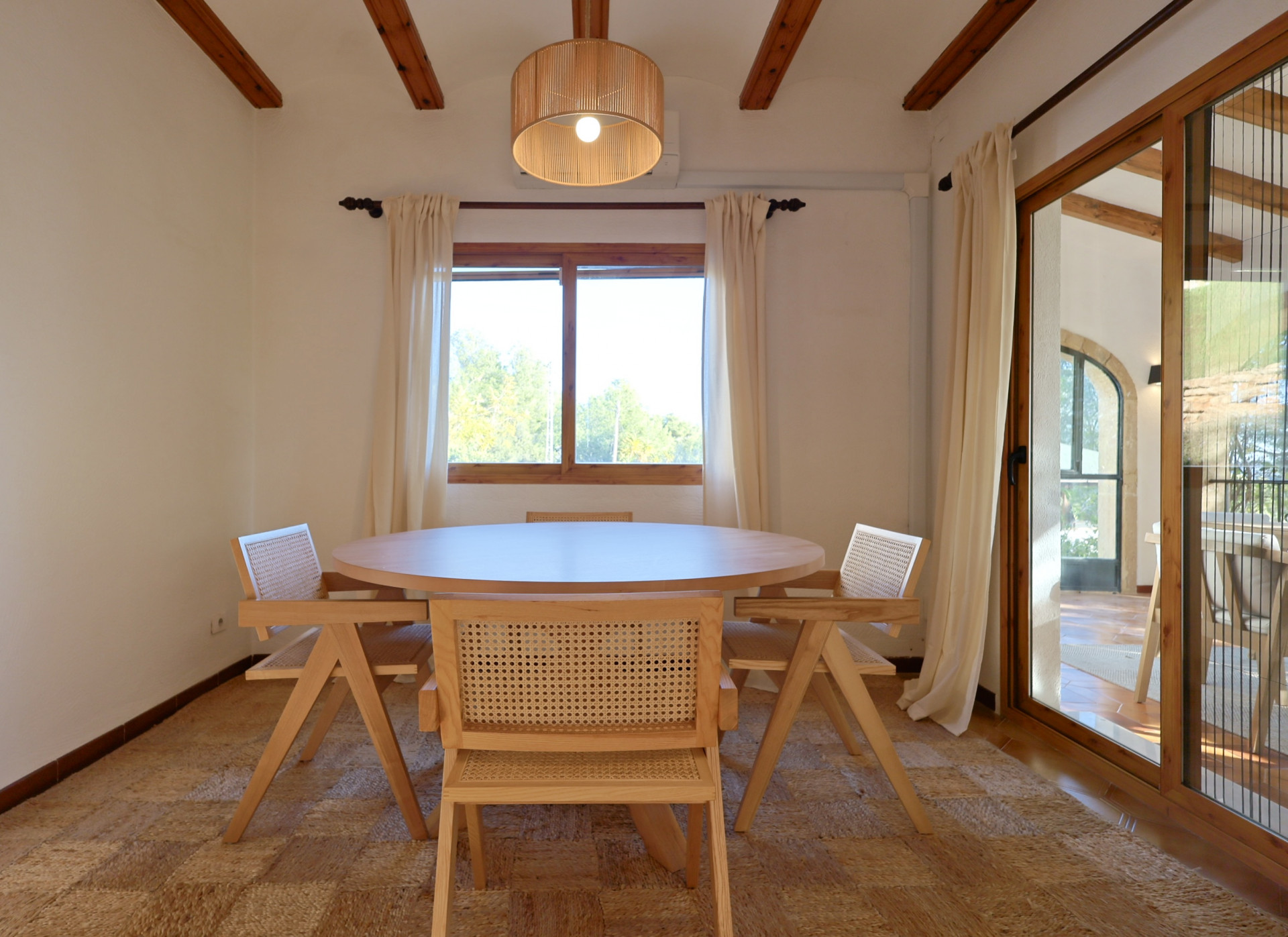 Comedor con mesa circular de madera, sillas de rejilla y alfombra de fibras naturales, bajo un techo de vigas vistas y lámpara de láminas de madera.