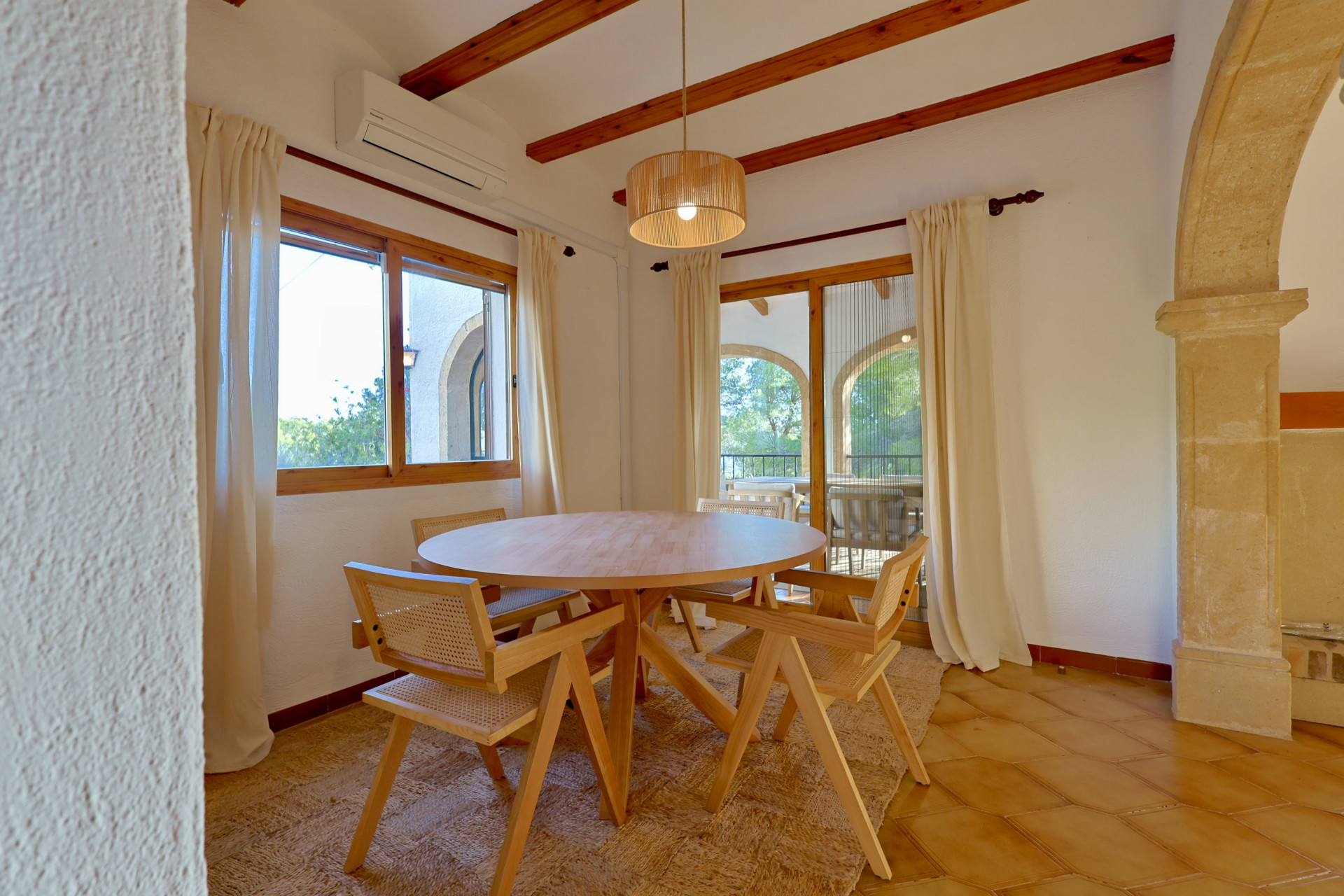Comedor con mesa redonda de madera y sillas de rejilla sobre suelo de terracota, vigas de madera a la vista y un arco de piedra natural.