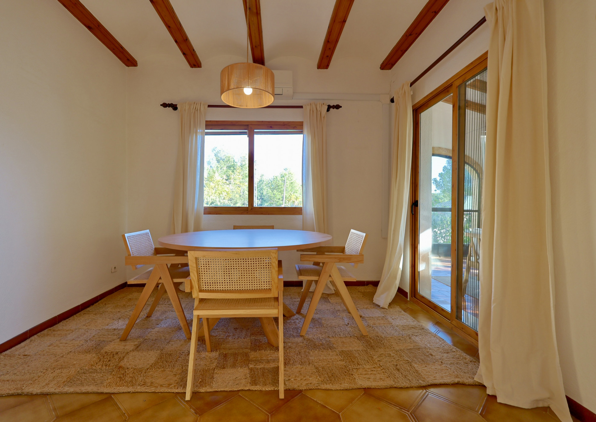 Comedor con vigas de madera expuestas, mesa redonda y sillas de rejilla sobre alfombra de fibras naturales. Incluye acceso directo a la terraza mediante puertas correderas.
