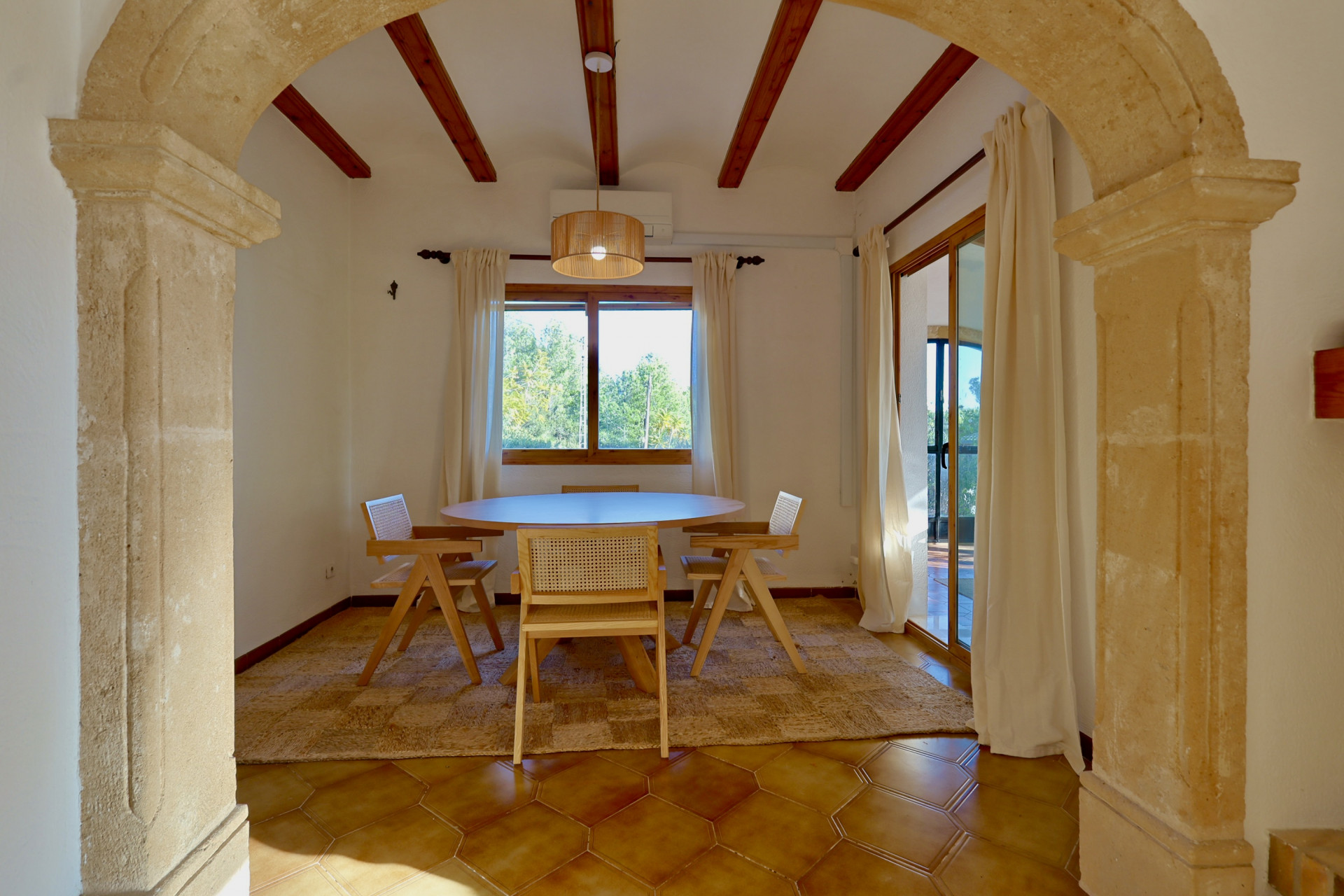 Comedor rústico enmarcado por un arco de piedra, con vigas de madera expuestas, suelos de terracota hexagonal y mobiliario de madera con detalles de ratán.