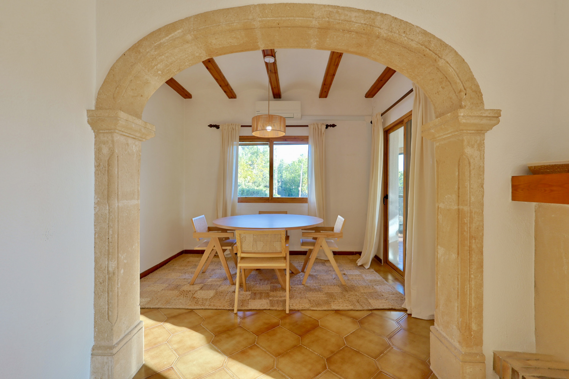 Comedor rústico enmarcado por un arco de piedra natural, con vigas de madera expuestas, suelos de terracota hexagonal y mobiliario de madera y ratán.