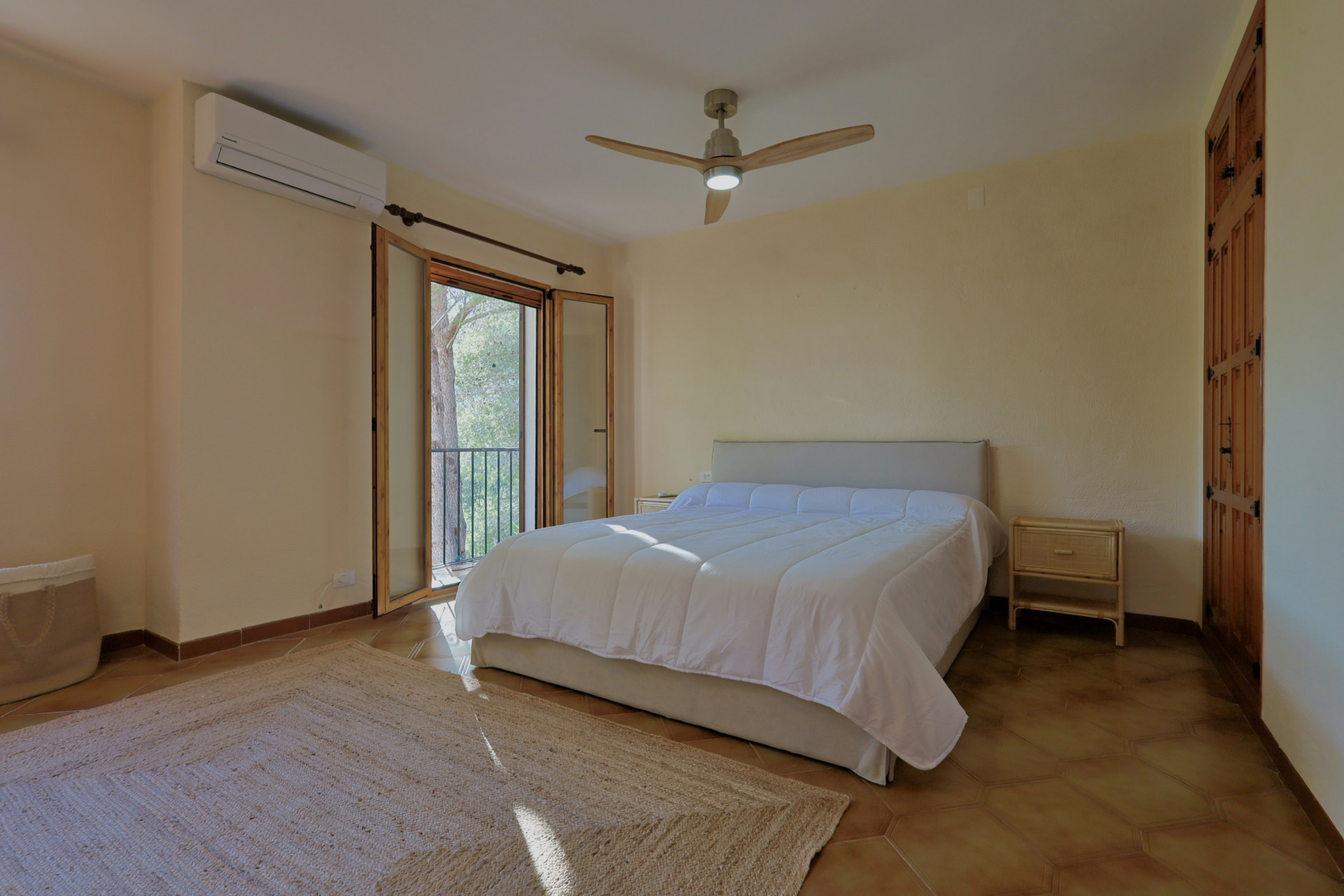 Dormitorio con suelos de terracota, cama doble y ventilador de techo de madera. Incluye puertas francesas hacia un balcón, armarios empotrados de madera y aire acondicionado.