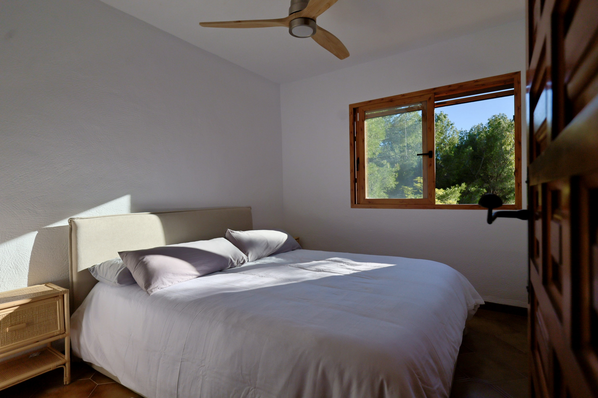 Dormitorio luminoso con ventilador de techo de madera, paredes blancas de yeso y ventana con vistas al bosque. Incluye cabecero tapizado y mesita de noche de mimbre.