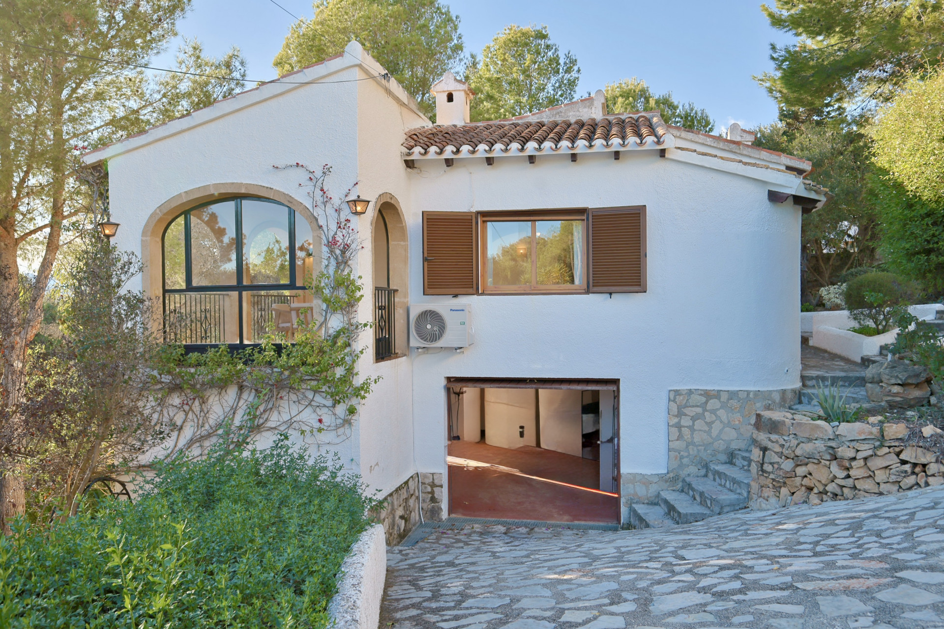Fachada de villa blanca con ventanal de arco en piedra, tejado de teja y garaje inferior integrado en un entorno natural con pinos y muros de piedra.