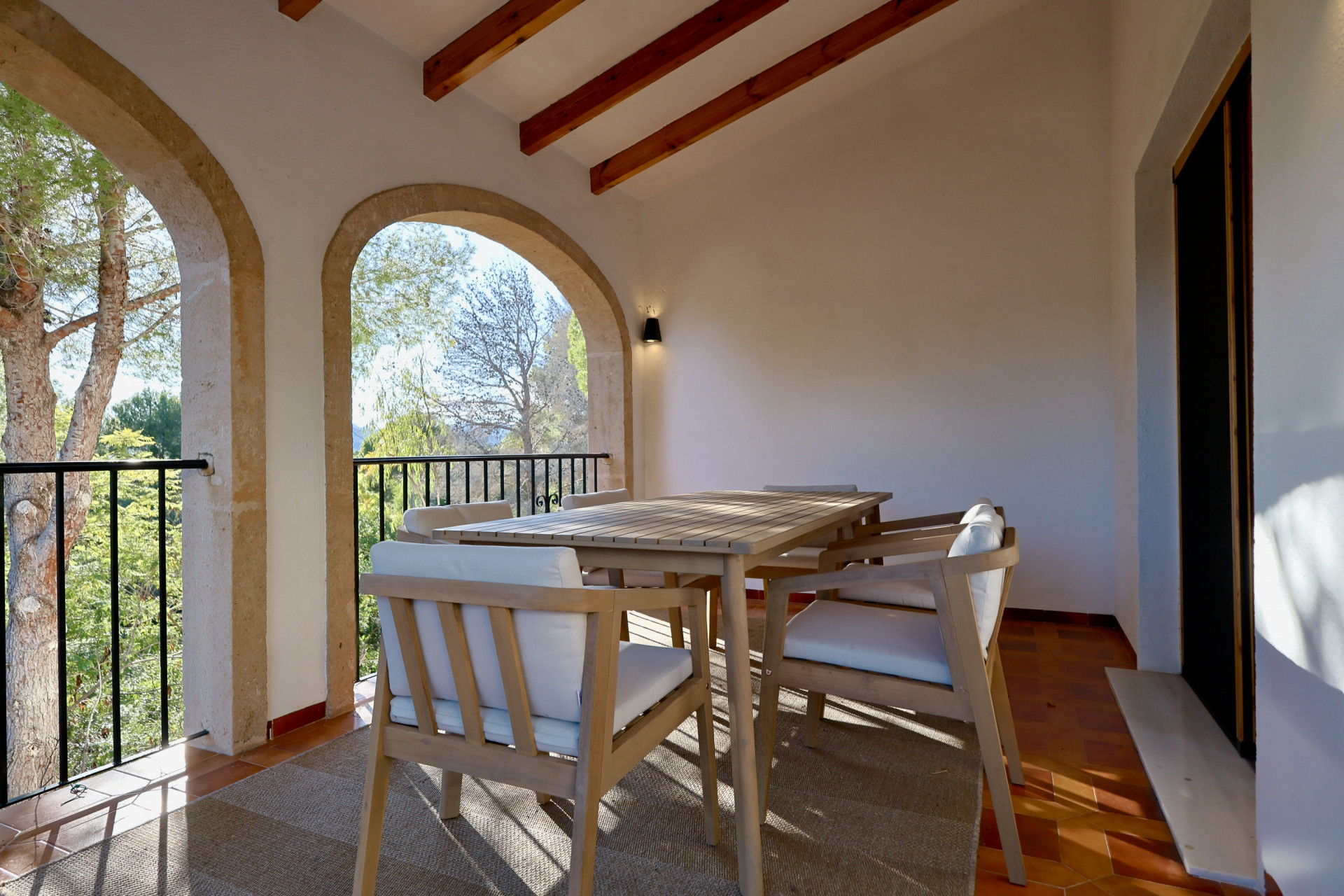 Porche cubierto con arcos de piedra tosca, vigas de madera a la vista y zona de comedor exterior con vistas al entorno arbolado.