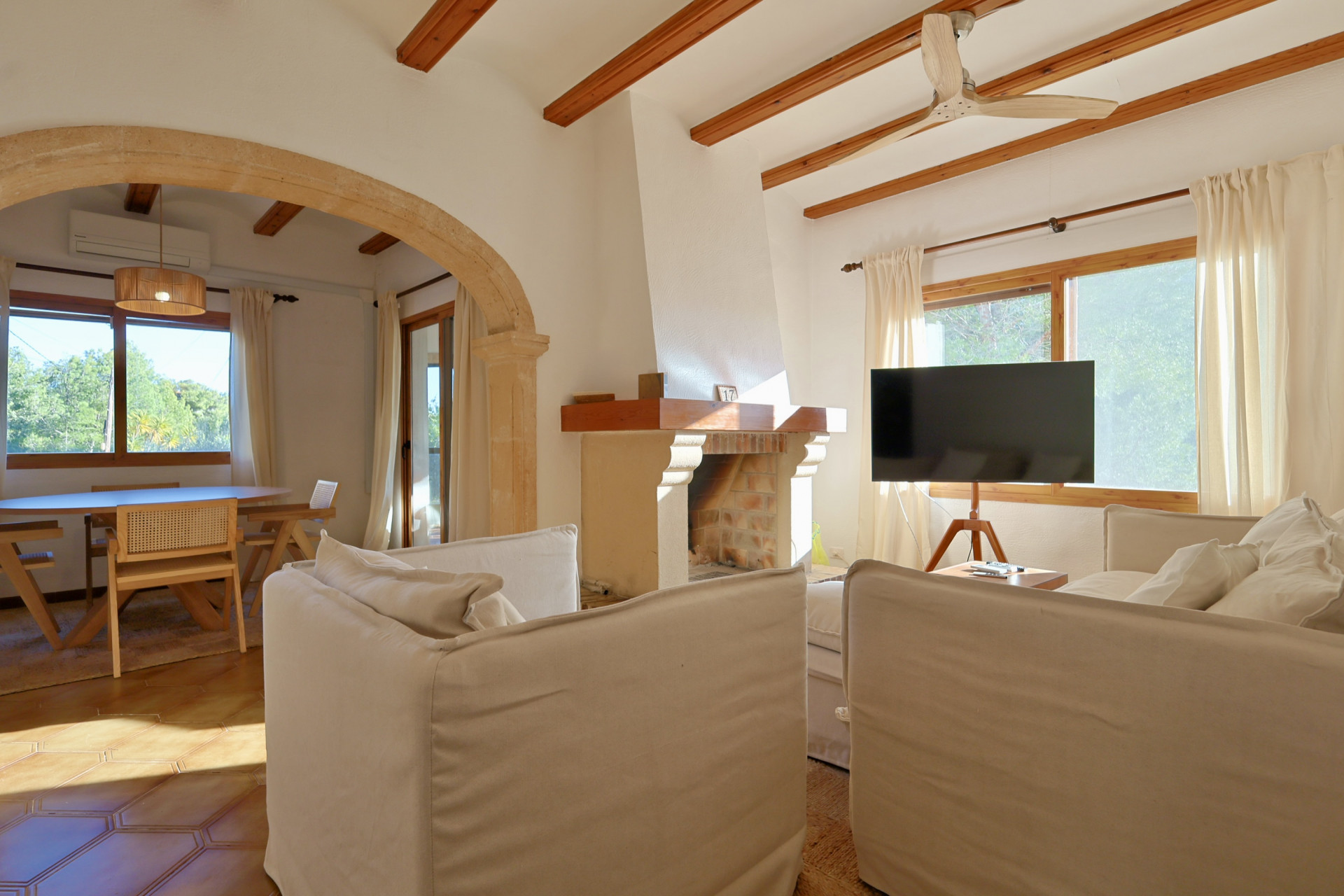 Salón con vigas de madera vistas, arco de piedra y chimenea de ladrillo. Dispone de sofás blancos y zona de comedor con suelo de baldosas de terracota.