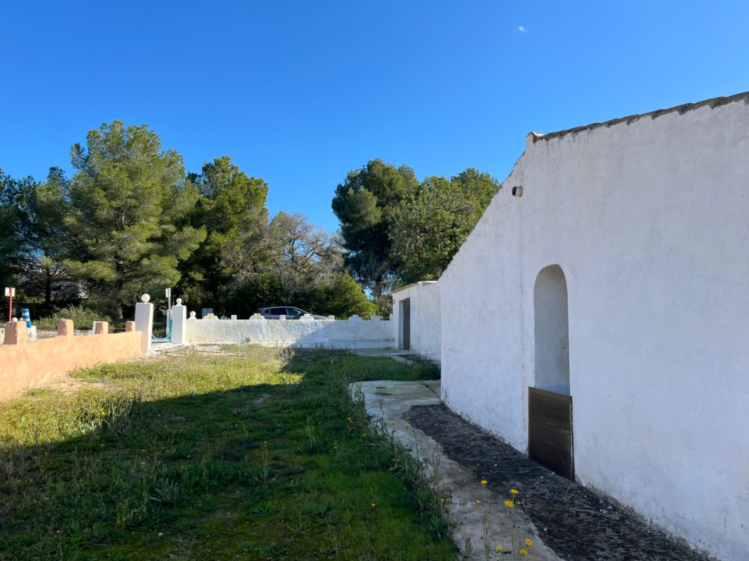 Fachada blanca de una finca tradicional en Jávea con nicho de arco y terreno natural, delimitada por muros blancos y pinos bajo un cielo despejado.