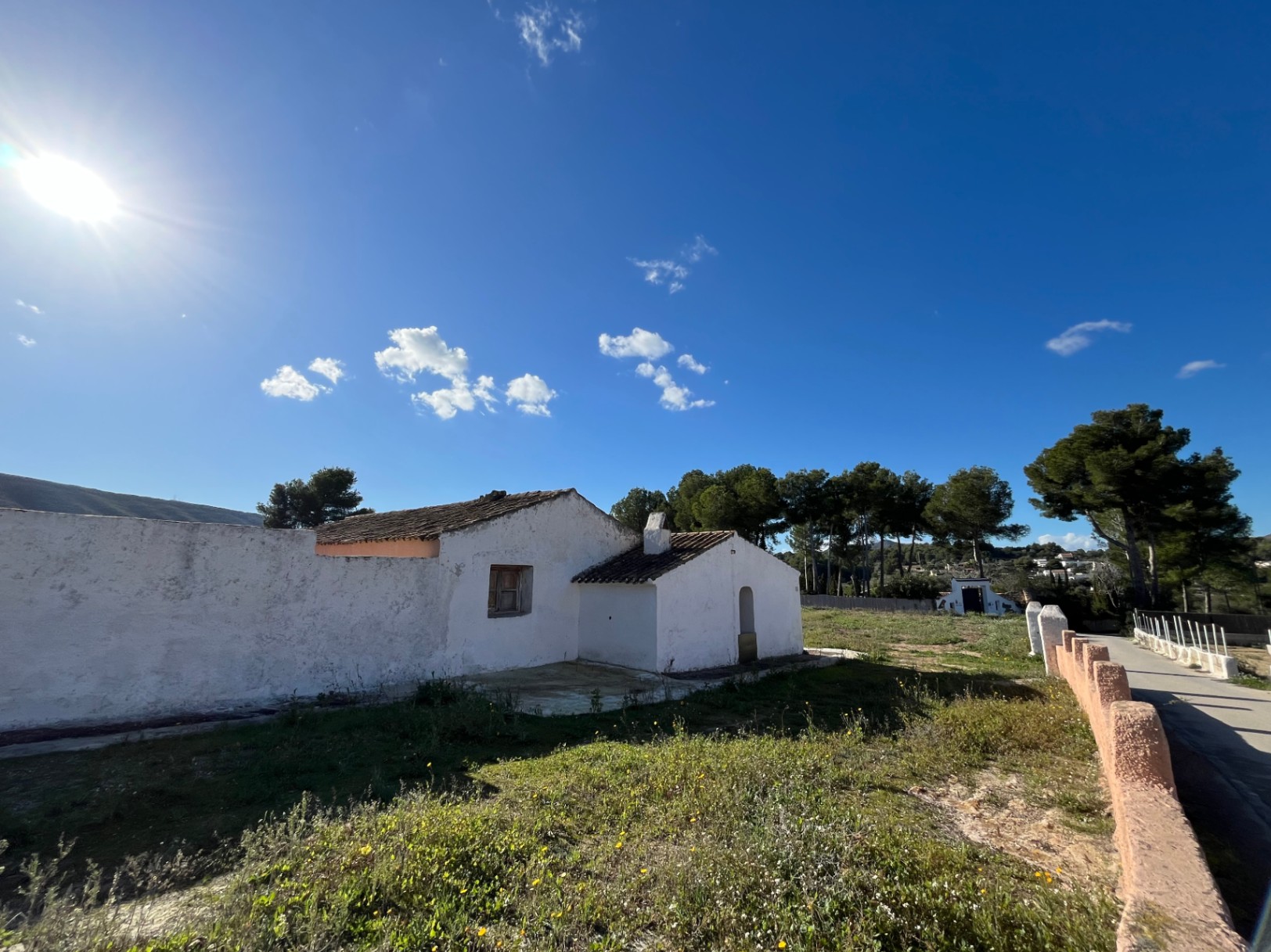 Fachada exterior de finca tradicional con muros blancos y tejado de teja, rodeada de terreno natural bajo un cielo despejado en Jávea.