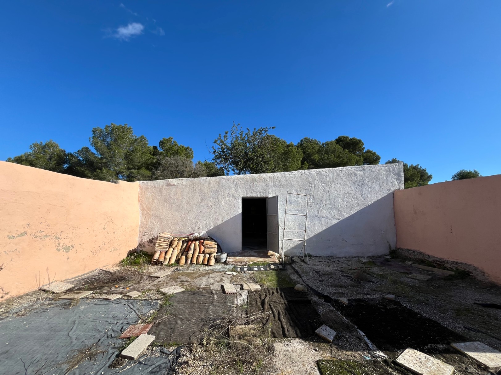 Patio cerrado con construcción auxiliar encalada, apilamiento de tejas cerámicas tradicionales y muros perimetrales de color terracota.
