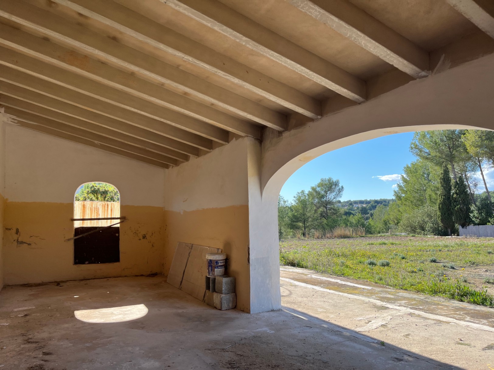 Porche en reforma con techo de vigas vistas y un gran arco que ofrece vistas despejadas al entorno rural y la vegetación mediterránea.